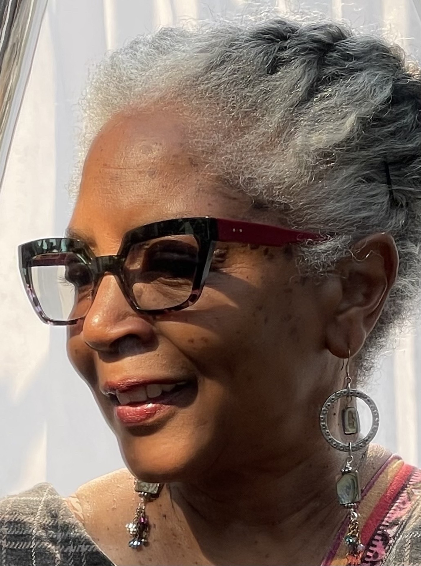 An older woman with gray hair styled in braids wears distinctive glasses and large earrings. She smiles warmly, exuding confidence and grace.